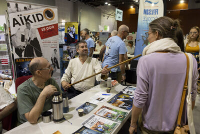 photo du stand de l'aïkido, deux hommes assis derrière, un homme en kimono tient un bâton qu'il montre à une femme devant le stand - Agrandir l'image 18 sur 68, fenêtre modale