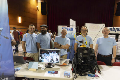 photo d'un stand à l'intérieur, cinq hommes avec un tee-shirt bleu sourient à l'objectif, un écran est posé sur la table avec une vue sous-marine, un équipement de plongée (bouteille et sac à dos) est posé sur la table aussi - Agrandir l'image 19 sur 68, fenêtre modale