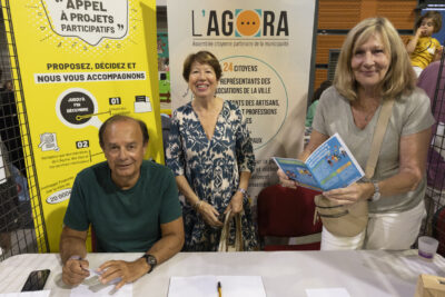 photo du stand de l'agora et l'appel à projets participatifs, deux femmes et un homme derrière sourient à l'objectif. Sur la table, il y a des dépliants jaunes sur l'appel à projets participatifs et des dépliants bleus à côté - Agrandir l'image 25 sur 68, fenêtre modale