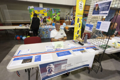 photo du stand du comité laïcité et république, un homme derrière le stand est assis et sourit à l'objectif - Agrandir l'image 26 sur 68, fenêtre modale