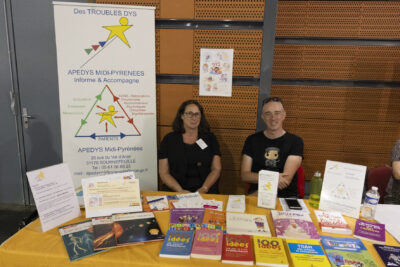 photo du stand Apedys, autour des troubles dys, sur la table, de nombreux livres, derrière la table un homme et une femme assis qui sourient à l'objectif - Agrandir l'image 27 sur 68, fenêtre modale