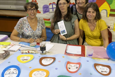 photo d'un stand lié à la petite enfance, derrière quatre femmes sourient à l'objectif, sur la table sont collés des visages dessinés avec des expressions différentes, et derrière elles une bâche colorée avec des photos réparties dessus - Agrandir l'image 29 sur 68, fenêtre modale