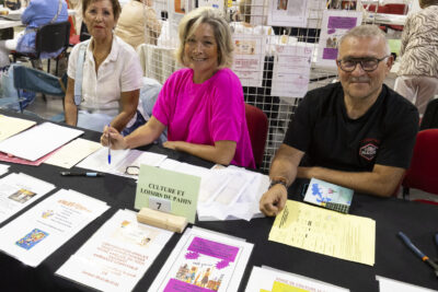 photo du stand de culture et loisirs de Pahin, deux femmes et un homme assis derrière sourient à l'objectif, des documents sont répartis sur la table - Agrandir l'image 30 sur 68, fenêtre modale