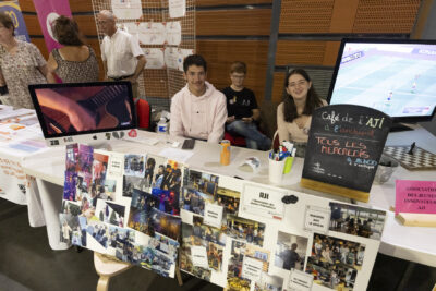 photo du stand de l'AJI à l'archipel, trois jeunes sont derrière la table. deux panneaux avec des collages photos sont accrochés devant le stand, il y a aussi un écran avec une vue rapprochée sur une main à la guitare - Agrandir l'image 31 sur 68, fenêtre modale