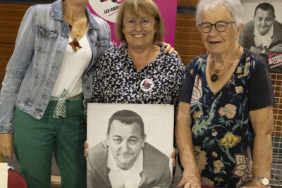 photo du stand des restaurants du coeur, trois femmes posent derrière en souriant et en tenant le portrait noir et blanc de Coluche - Agrandir l'image 32 sur 68, fenêtre modale