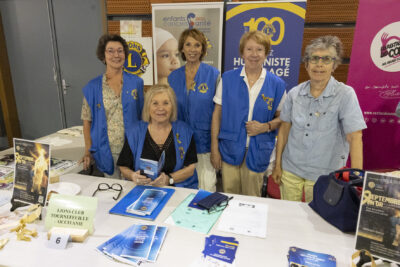 photo du stand du lions club tournefeuille occitanie, cinq femmes posent derrière, quatre portent un gilet bleu avec le logo bleu et jaune de l'association, sur la table divers supports papiers sont répartis - Agrandir l'image 33 sur 68, fenêtre modale