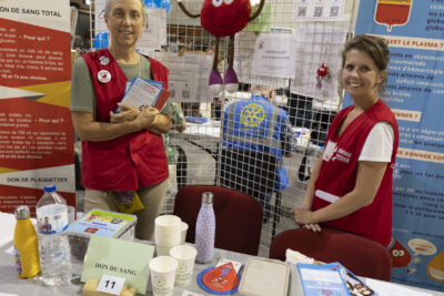 photo du don du sang avec deux femme sderrière qui portent un gilet rouge et sourient à l'objectif, la mascotte (une goutte de sang rouge avec un sourire et des yeux façon dessin animé) entre elles, et divers supports répartis sur la table - Agrandir l'image 34 sur 68, fenêtre modale