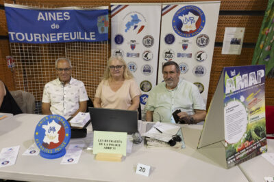 photo du stand de l'association des retraités de la Police, derrière le stand deux hommes et une femme assis qui sourient à l'objectif - Agrandir l'image 35 sur 68, fenêtre modale