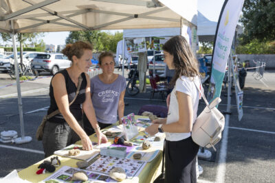 photo d'un stand associatif en extérieur, deux femmes derrière le stand échangent avec une jeune femme - Agrandir l'image 5 sur 68, fenêtre modale