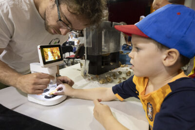 photo d'un stand, un jeune homme montre à un petit garçon un minéral sous un microscope avec un écran - Agrandir l'image 45 sur 68, fenêtre modale