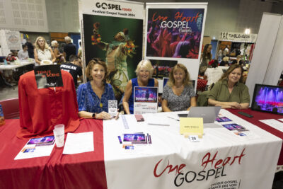 photo du stand de one heart gospel avec quatre femmes derrière le stand qui sourient à l'objectif en montrant une petite affiche sur laquelle il est écrit "rejoignez-nous", usr le stand, des suppports papiers et une tenue de scène rouge sur un mannequin - Agrandir l'image 46 sur 68, fenêtre modale