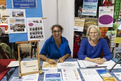 photo du stand de Choeur méditerranée, deux femmes habillées en bleu sourient à l'objectif, divers supports bleus sont éparpillés sur la table - Agrandir l'image 47 sur 68, fenêtre modale