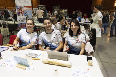 photo du stand de badminton, trois personnes derrière portent un maillot blanc et bleu, et montrent leur raquette en souriant à l'objectif - Agrandir l'image 50 sur 68, fenêtre modale