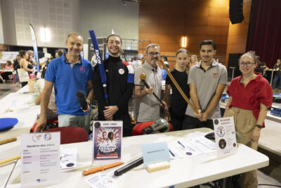 photo du stand de karaté genbudo, six personnes derrière le stand sourient et tiennent dans leur main des sortes de sabres en bois - Agrandir l'image 52 sur 68, fenêtre modale