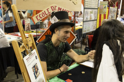 photo du stand d'Espantous, école de magie, avec un magicien en tenue qui montre un tour à une femme penchée sur le stand - Agrandir l'image 59 sur 68, fenêtre modale