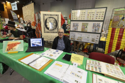 photo du stand de Tournephil, un homme assis regarde l'objectif, plein de timbres sont disposés sur la table et sur des panneaux derrière - Agrandir l'image 61 sur 68, fenêtre modale