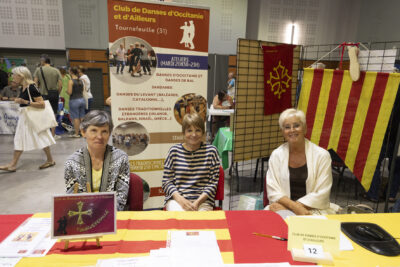 photo du stand de club de danses d'Occitanie et d'ailleurs, trois femmes assises derrière une table aux couleurs jaune et rouge de l'Occitanie - Agrandir l'image 63 sur 68, fenêtre modale