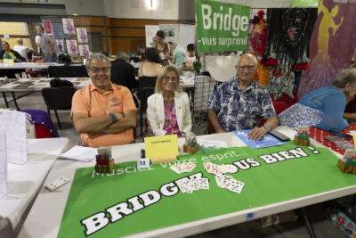 photo du stand de tournebridge, trois personnes sourient à l'objectif assises derrière la table, sur laquelle il y a un tapis vert avec écrit "bridgez vous bien" et des cartes disposées par couleurs - Agrandir l'image 65 sur 68, fenêtre modale