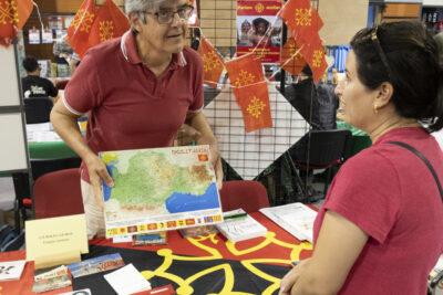 photo d'un homme qui échange avec une femme à un stand, il lui montre une carte relief d'Occitanie, sur la table on voit la croix occitane - Agrandir l'image 67 sur 68, fenêtre modale