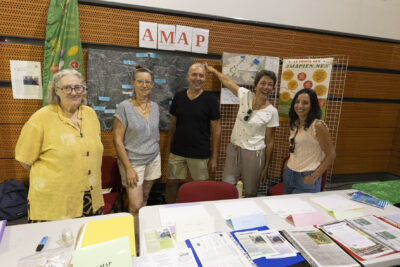 photo du stand de l'amap avec cinq personnes derrière qui sourient à l'objectif, une femme montre du doigt quatre feuilles accrochées au mur derrière sur lesquelles il est écrit AMAP - Agrandir l'image 68 sur 68, fenêtre modale