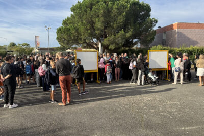 Photo des familles rassemblées devant deux tableaux blancs dans une cour, sous un ciel bleu - Agrandir l'image 14 sur 80, fenêtre modale
