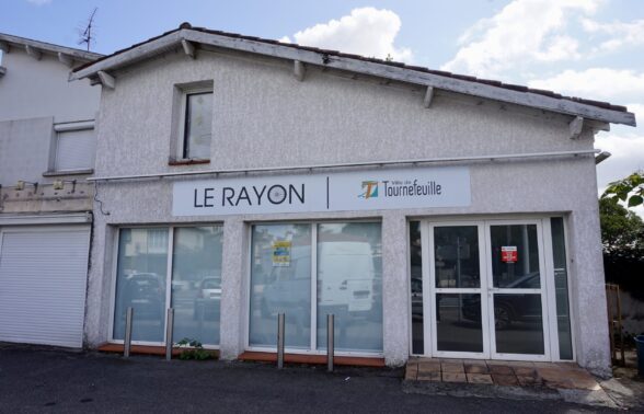 photo de la devanture du local du Rayon, on voit une façade blanche avec l'enseigne Ville de Tournefeuille Le rayon, et au rez-de-chaussée une porte vitrée et des vitres fumées