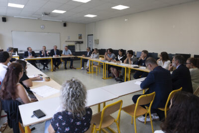 photo d'une réunion dans une salle de classe, vue d'ensemble de la trentaine de personnes réunies, certaines en costumes - Agrandir l'image 56 sur 80, fenêtre modale