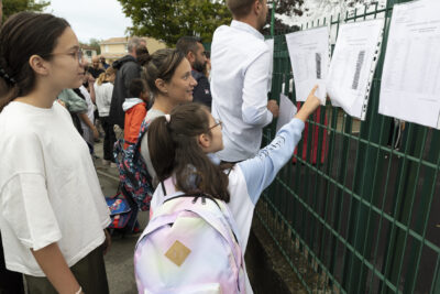 des élèves rassemblés devant les listes de classes accrochées aux grilles vertes de la cou, une jeune fille le doit posé sur une des listes - Agrandir l'image 47 sur 80, fenêtre modale