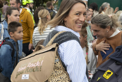 une femme avec un cartable au dos se retourne pour sourire à l'objectif, de nombreuses familles réunies derrière elle - Agrandir l'image 48 sur 80, fenêtre modale