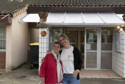 deux femmes posent devant l'entrée de l'école, sous une ombrière en forme de feuille, l'une tient l'autre par l'épaule, elles sourient - Agrandir l'image 51 sur 80, fenêtre modale