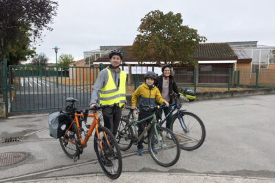 un homme, un enfant et une femme debout à côté de leurs vélos, devant la grille d'une cour d'école, le ciel gris - Agrandir l'image 52 sur 80, fenêtre modale