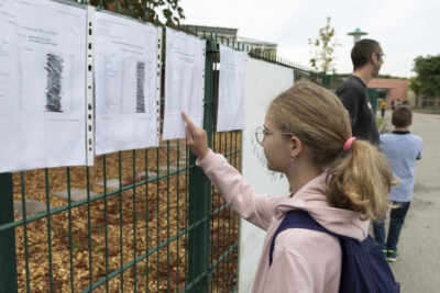 une jeune fille regarde une liste accrochée à la grille verte d'une cour d'école, le doigt sur la feuille pour y trouver son nom - Agrandir l'image 8 sur 80, fenêtre modale