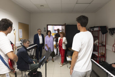 photo dans une salle de musique, trois jeunes, un en train de jouer de la guitare électrique, pendant qu'une femme en tenue rouge fait la visite au Maire et élus - Agrandir l'image 57 sur 80, fenêtre modale