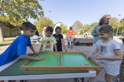 Photo paysage de deux enfants qui jouent à un jeu en bois posé sur une table, devant deux autres enfants qui regardent, sur une place - Agrandir l'image 4 sur 16, fenêtre modale