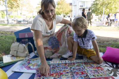 Photo d'une femme et d'un petit garçon, assis par terre sur un tapis extérieur, à regarder une sorte de plateau avec plein de personnages dessinés. Elle lui montre du doigt quelque chose. En arrière-plan, la place ensoleillée - Agrandir l'image 5 sur 16, fenêtre modale