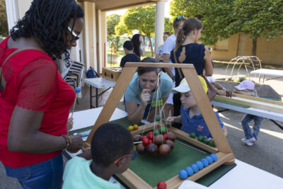 Photo de deux enfants qui jouent à un jeu en bois posé sur une table, deux femmes à leurs côtés en train de regarder le jeu, en extérieur - Agrandir l'image 7 sur 16, fenêtre modale