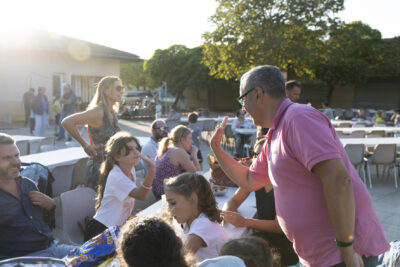 Photo paysage du Maire qui s'apprête à taper dans la main d'une petite fille à table avec d'autres enfants et adultes, tandis qu'il fait beau sur la place et qu'on peut voir d'autres tables positionnées en arrière-plan - Agrandir l'image 8 sur 16, fenêtre modale