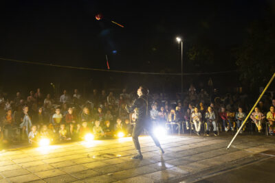Photo d'un spectacle nocturne, un homme au centre de la photo qui jongle avec un diabolo devant de nombreuses familles rassemblées, des enfants assis au premier rang - Agrandir l'image 12 sur 16, fenêtre modale