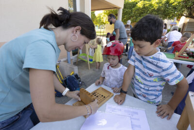 Photo paysage de deux enfants et une adulte, l'adulte montre un petit jeu en bois aux enfants, sur une table en extérieur. On voit d'autres enfants derrière eux jouer avec d'autres jeux - Agrandir l'image 2 sur 16, fenêtre modale