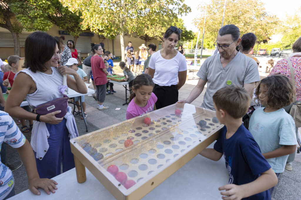 Photo paysage d'enfants qui jouent à un jeu en bois posé sur une table, des adultes souriants autour d'eux, sur une place ensoleillée et ombragée, avec d'autres familles en arrière-plan - Agrandir l'image, fenêtre modale