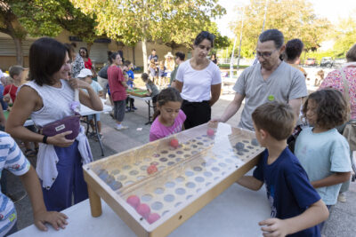 Photo paysage d'enfants qui jouent à un jeu en bois posé sur une table, des adultes souriants autour d'eux, sur une place ensoleillée et ombragée, avec d'autres familles en arrière-plan - Agrandir l'image 3 sur 16, fenêtre modale