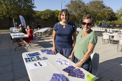Photo de deux femmes debout derrière une table, sur laquelle des dépliants sont disposés. Elle sourient à l'objectif - Agrandir l'image 15 sur 16, fenêtre modale