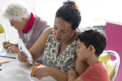 Photo d'une femme et d'un petit garçon, elle découpe une forme dans une feuille en papier sous les yeux du garçon - Agrandir l'image 13 sur 21, fenêtre modale