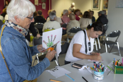 Photo d'une femme qui découpe une main dessinée sur une feuille blanche, on voit du monde à l'arrière-plan assis sur des chaises - Agrandir l'image 19 sur 21, fenêtre modale