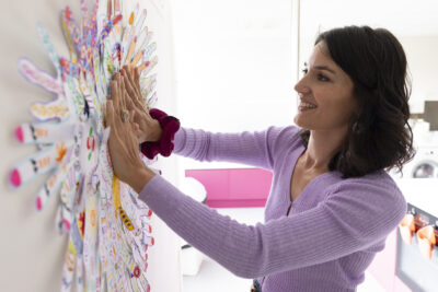 Photo d'une femme qui a les mains plaquées contre un mur sur lequel sont accrochées plein de formes de main qui forment une sorte de fleur - Agrandir l'image 11 sur 21, fenêtre modale