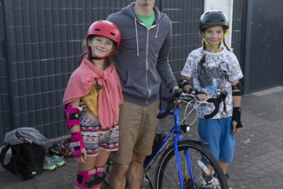 Photo d'un père avec ses deux enfants, il se tient à côté de son vélo et le tient par le guidon, de chaque côté un de ses enfants, tous deux à roller - Agrandir l'image 19 sur 80, fenêtre modale