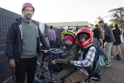 Photo d'un homme avec un casque vélo rose, qui tient son vélo cargo à l'arrière duquel deux enfants sont installés, les trois regardent l'objectif - Agrandir l'image 4 sur 80, fenêtre modale