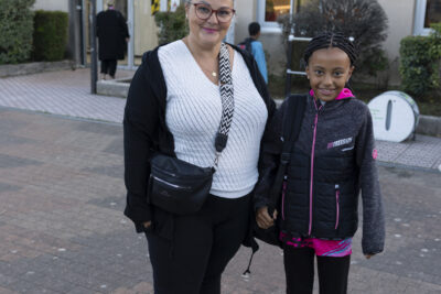Photo d'une femme debout avec sa fille, les deux souriantes, devant l'entrée du bâtiment de l'école - Agrandir l'image 20 sur 80, fenêtre modale