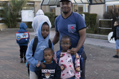 Photo d'un père avec ses trois enfants devant l'entrée de l'école, tout sourire - Agrandir l'image 24 sur 80, fenêtre modale