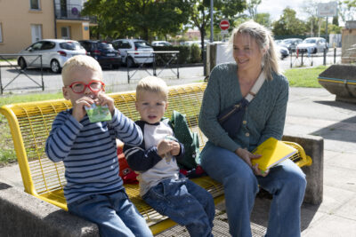 une femme et deux enfants assis sur un banc jaune au soleil, un des enfants mange une pompote - Agrandir l'image 65 sur 80, fenêtre modale